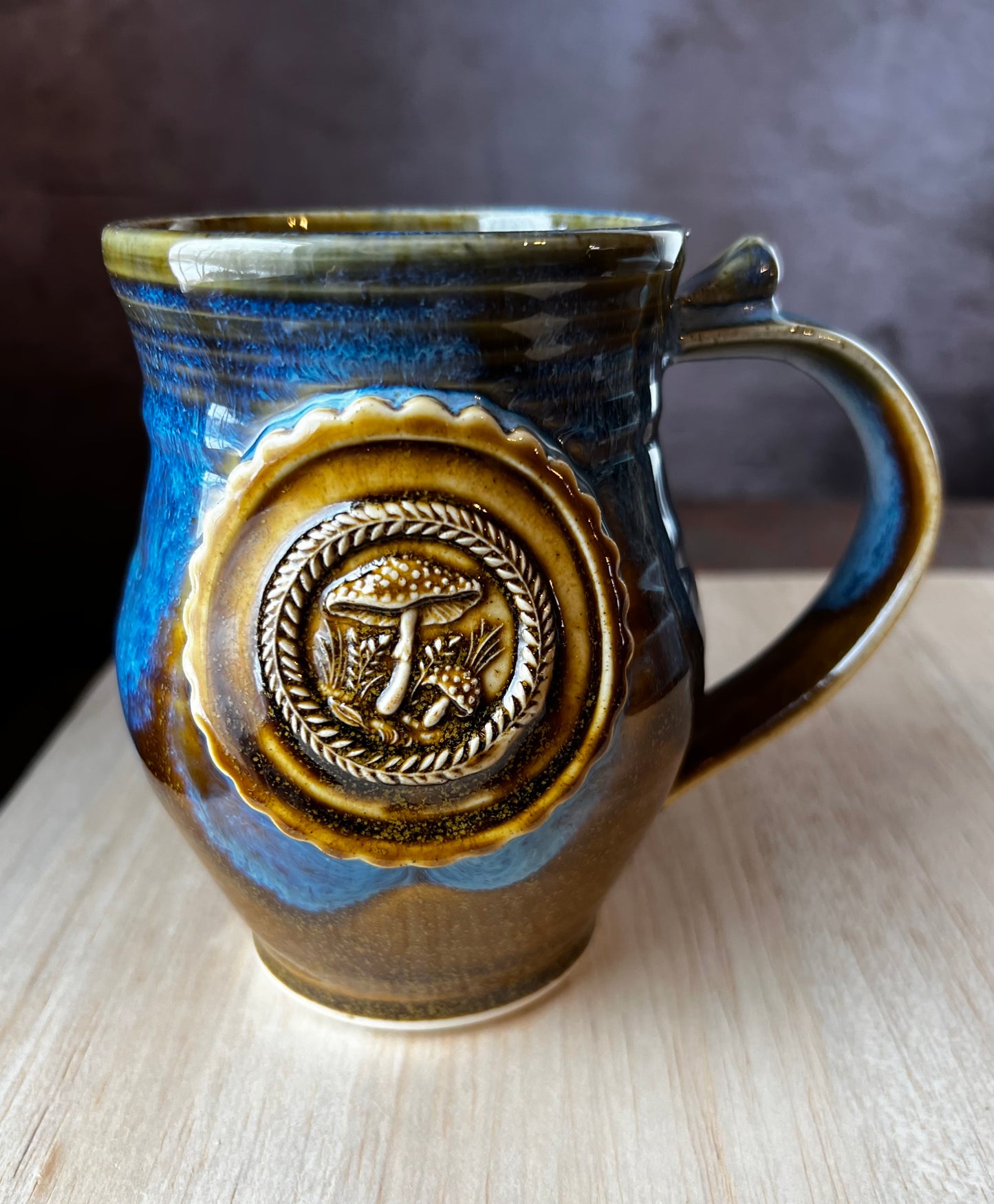 A left-handed ceramic mug in brown and blue with a medallion featuring 2 Amanita mushrooms