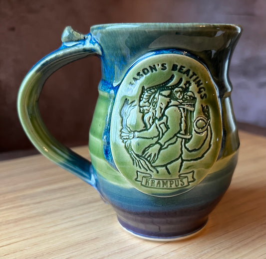 A ceramic mug with a green and blue color scheme, featuring a mythical beast design and the text 'Season's Beatings'.