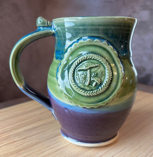 A ceramic mug with a green, blue, and purple color scheme, featuring a whimsical medallion adorned with amanita mushrooms.