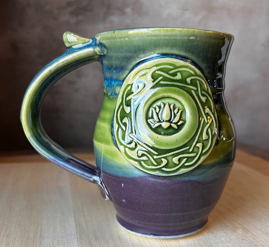A handcrafted ceramic mug with a lotus medallion and Celtic knotwork in black, gold, and green colors. Perfect for after yoga tea relaxaion.