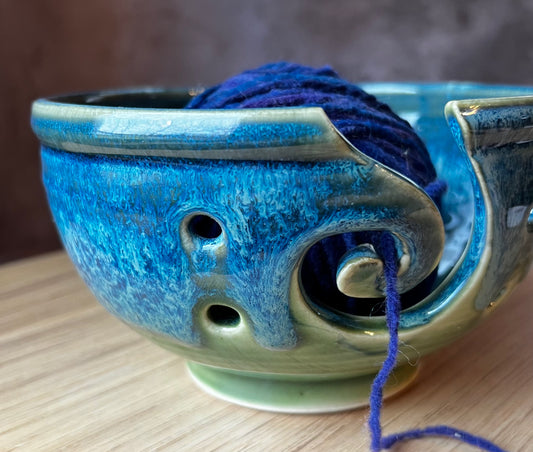 A handmade ceramic yarn bowl with a spiral design, holding a skein of blue yarn, placed on a wooden surface.