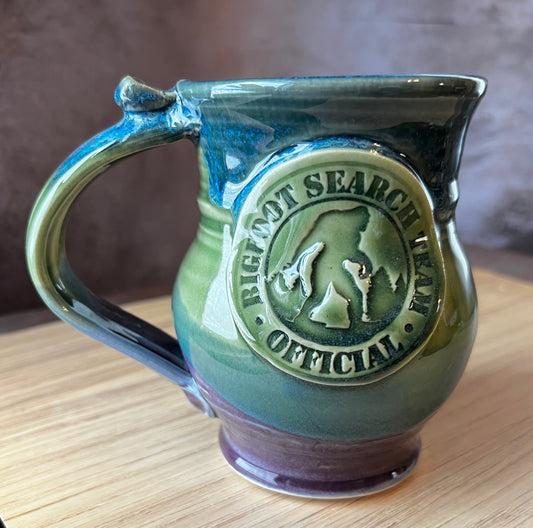 A ceramic mug with a green, blue, and purple color scheme, featuring a distinctive Bigfoot silhouette medallion on the front.