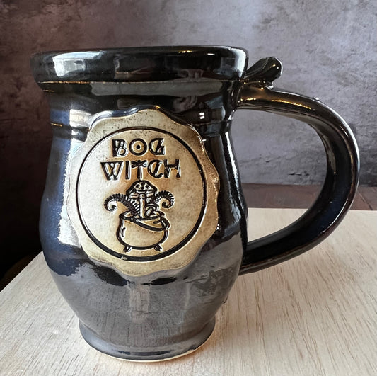A left-handed black mug with a medallion featuring a cauldron with mushrooms, and plants poking out the top the words "Bog Witch" are above the cauldron.
