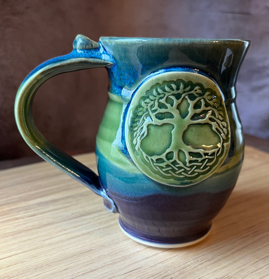 A multicolored ceramic mug with a handle, featuring an intricate Tree of Life motif in green on a background that includes shades of blue and green.