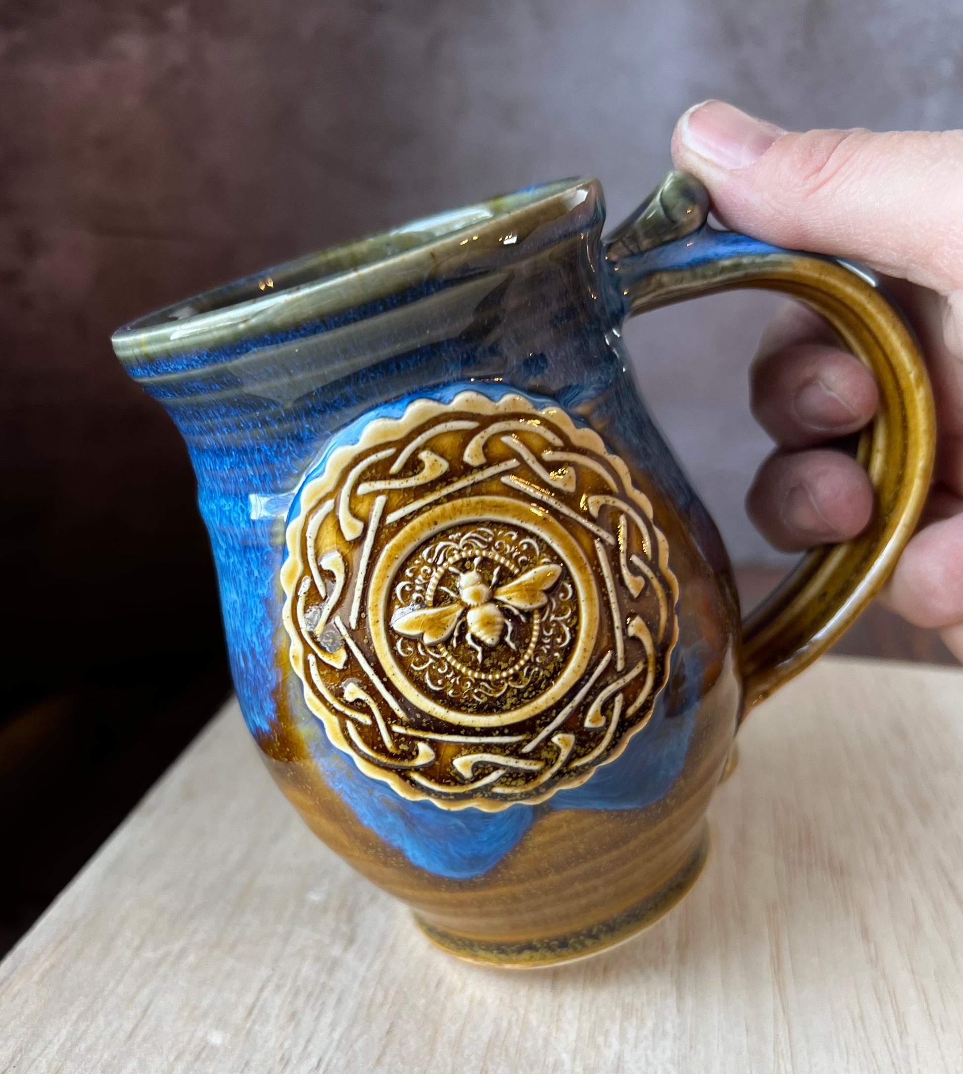 Left-handed ceramic mug in brown and blue with a medallion featuring a honeybee within a circle of Celtic knotwork