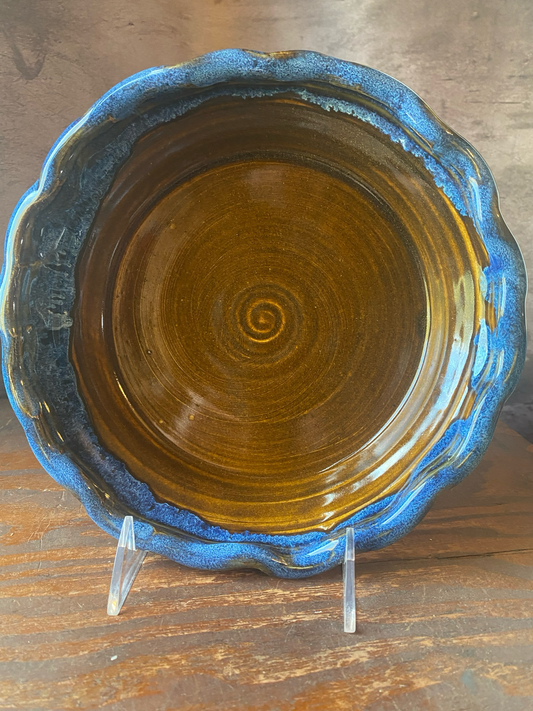 A handmade stoneware pie baker with a blue rim and a ruffled edge and brown glazed center. 