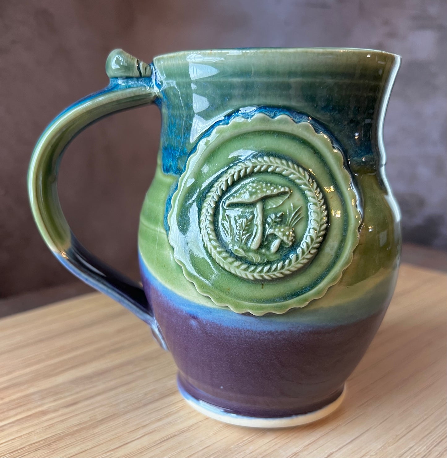 A ceramic mug with a green, blue, and purple color scheme, featuring a whimsical medallion adorned with amanita mushrooms.