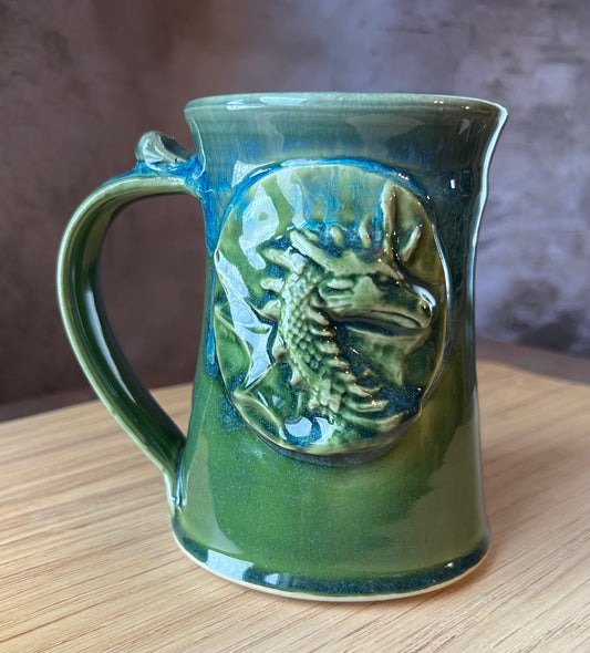 A ceramic tankard with a green and blue color scheme, featuring a sculptural dragon design on the side.