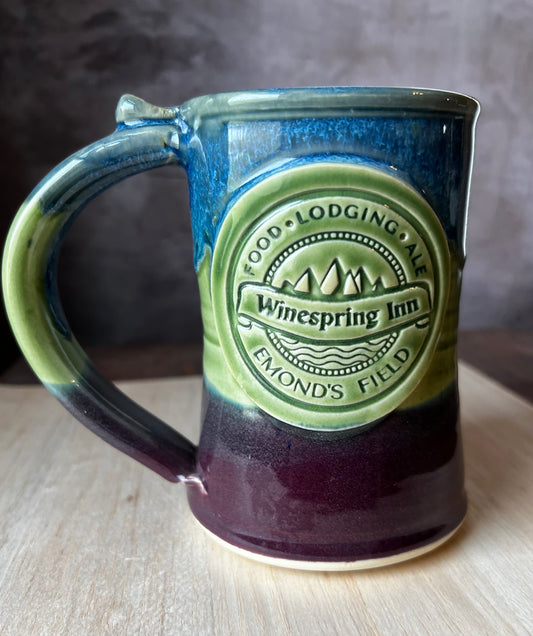 A ceramic mug with a green and blue color scheme, featuring a detailed medallion of the Winespring Inn emblem, and text reading 'Winespring Inn - Food. Lodging.