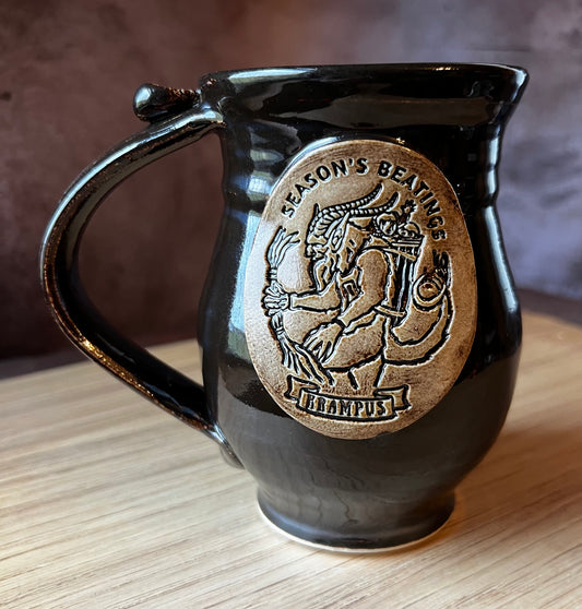 A black ceramic mug with a mythical beast design, specifically a Krampus, on the front. 