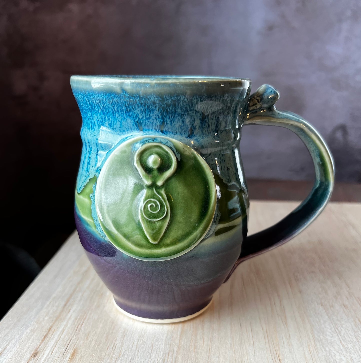 Left-handed mug in purple, green and blue featuring a medallion with a depiction of a traditional goddess image hands raised over her head with a spiral leading from her heart to her rounded belly