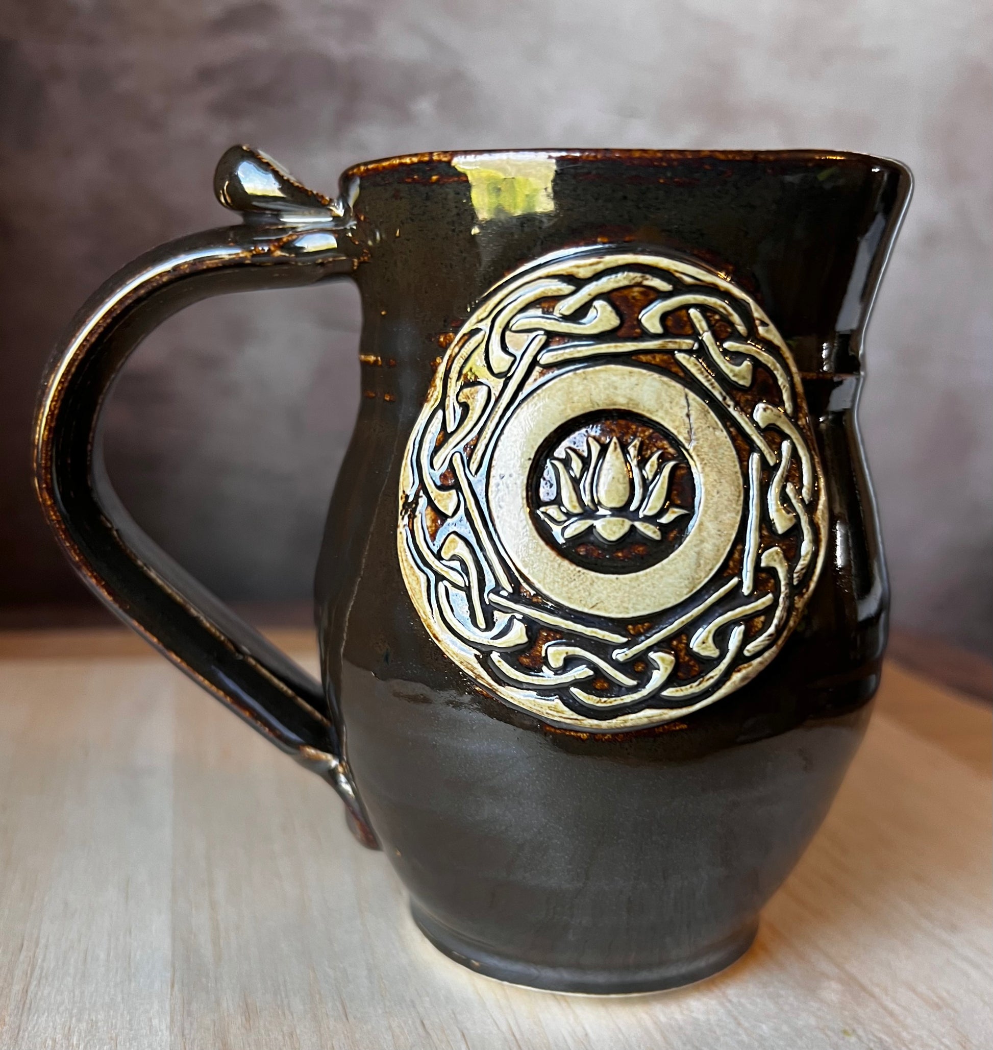 A handcrafted ceramic mug with a lotus medallion and Celtic knotwork in black, gold, and green colors.