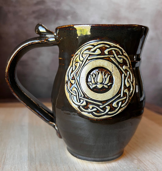 A handcrafted ceramic mug with a lotus medallion and Celtic knotwork in black, gold, and green colors.