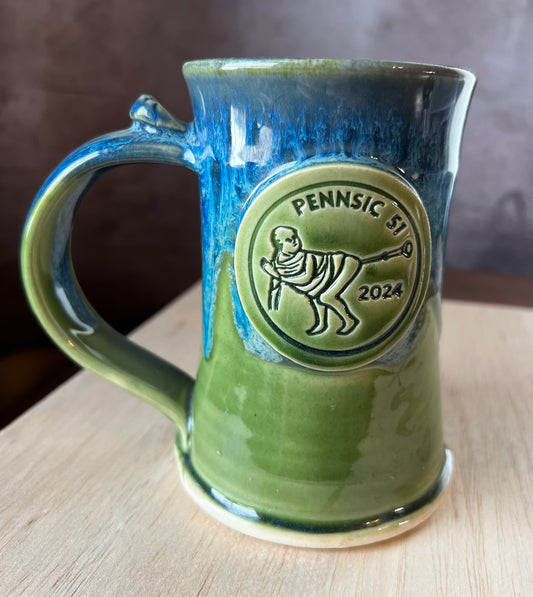 Green and blue ceramic mug with a decorative medallion featuring a medieval illumination of a monk blowing a trumpet with his butt. It is a commemorative mug or Pennsic 51 (2024) an event put on by the SCA, The Society for Creative Anachronism
