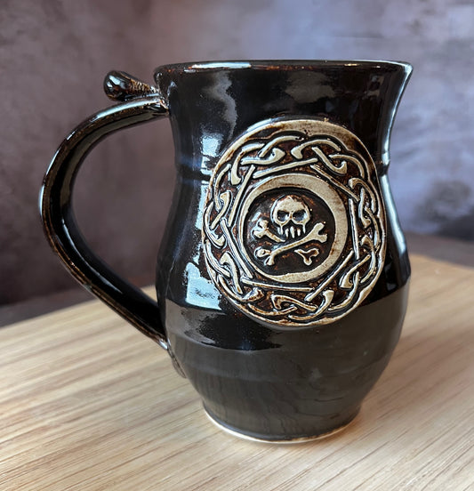 Black pottery mug with a celtic knot border surrounding a skull and cross bones.