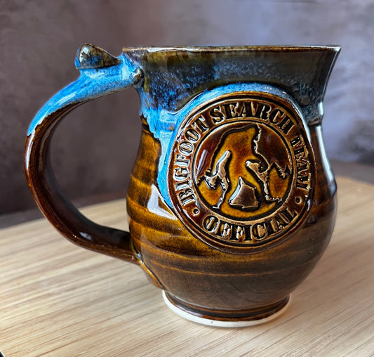 A ceramic mug with a blue handle and a distinctive Bigfoot silhouette medallion in the center.
