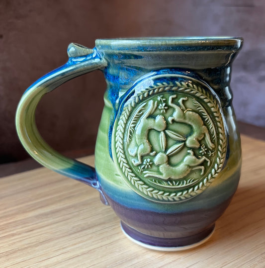 A handmade ceramic mug with a green, blue, and purple color scheme, featuring an intricate triple hare design in the center.