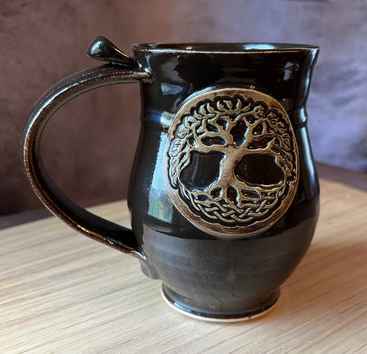 A black ceramic mug with a handle and a Tree of Life design in the center.