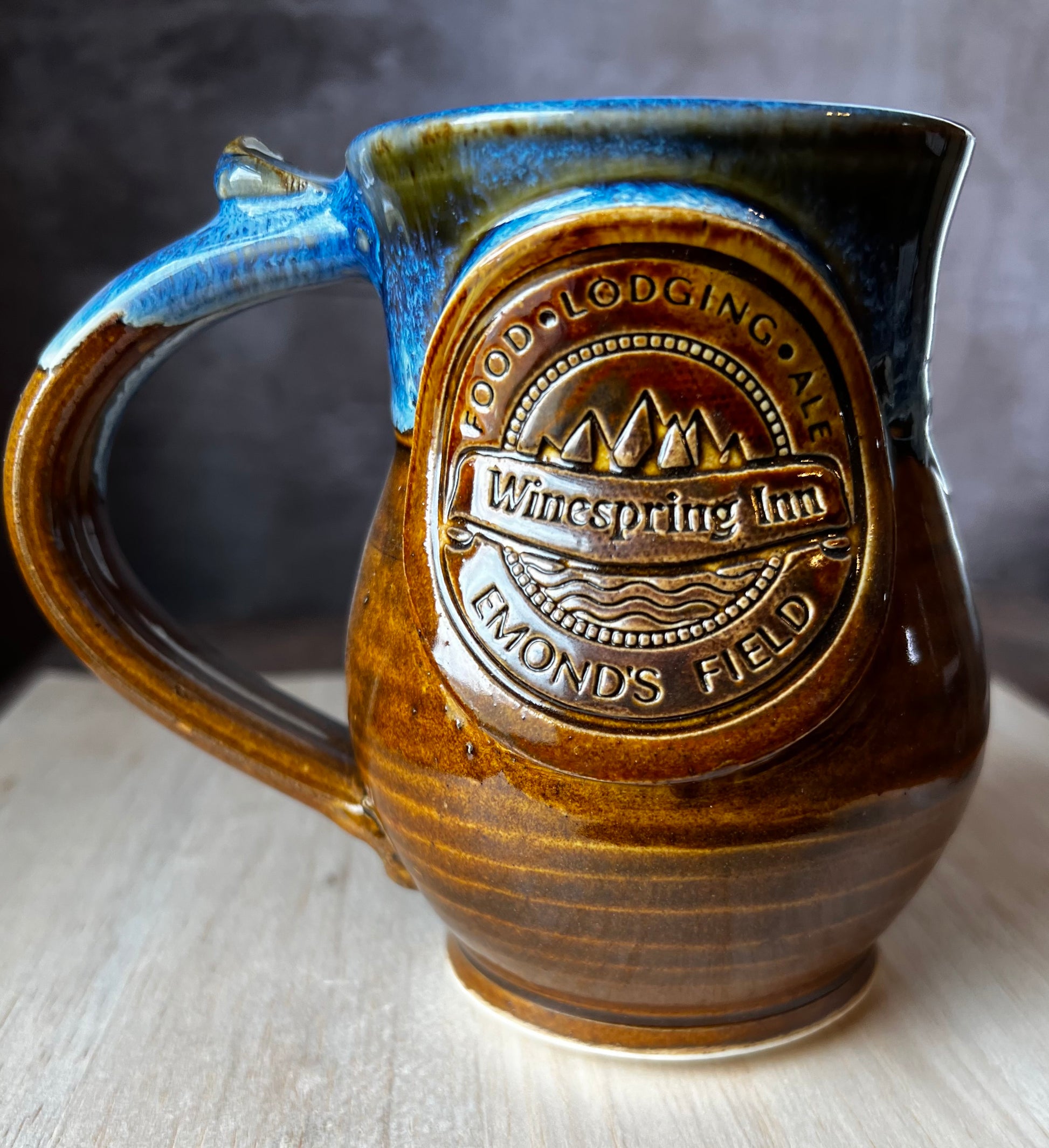 A ceramic mug with a rustic design, featuring a detailed medallion of the Winespring Inn on the front medallion