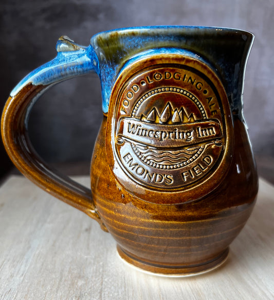A ceramic mug with a rustic design, featuring a detailed medallion of the Winespring Inn on the front medallion