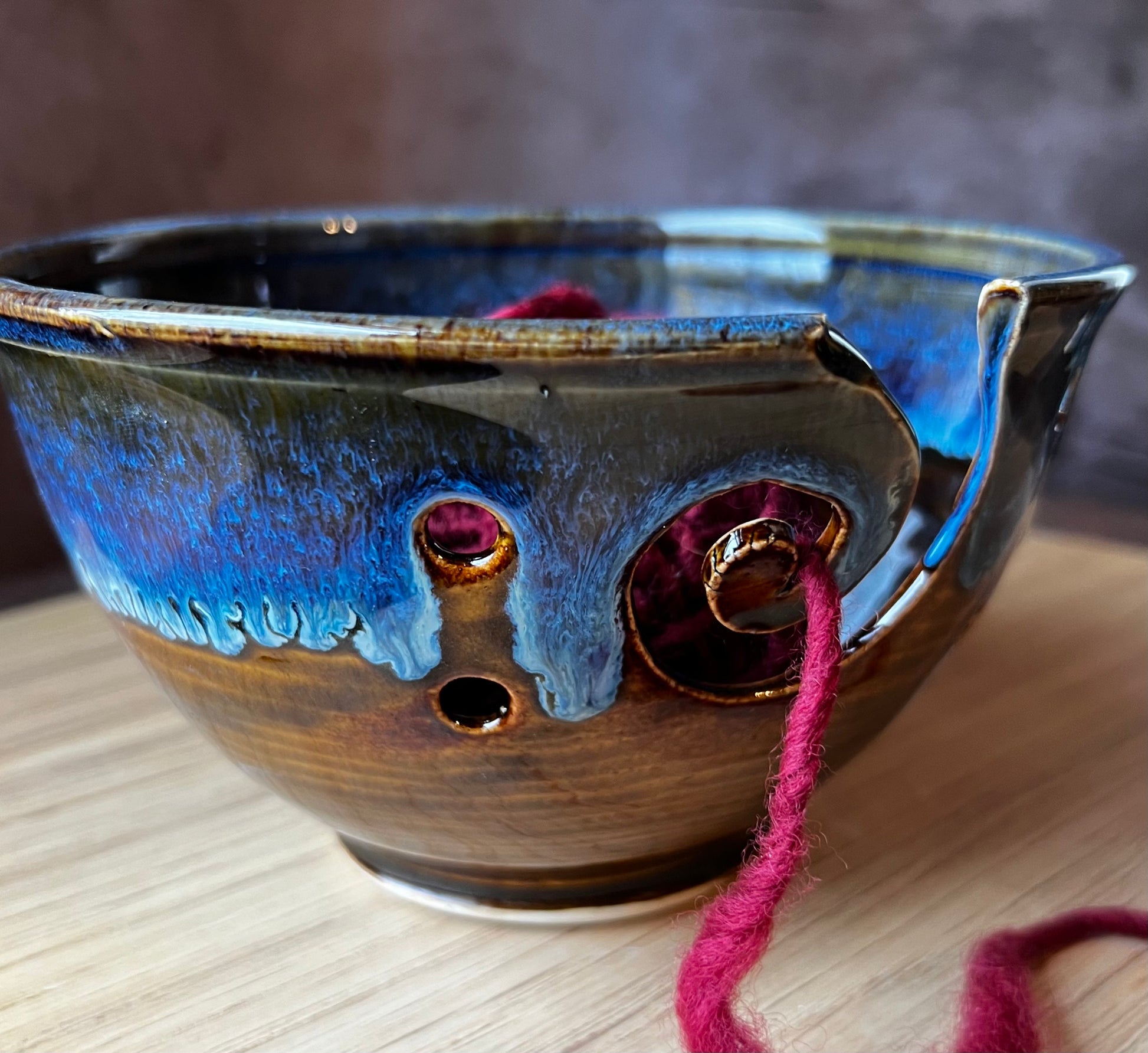 A handmade ceramic yarn bowl with blue and brown colors and a red yarn ball inside. The bowl has a handle and extra holes for managing yarn.