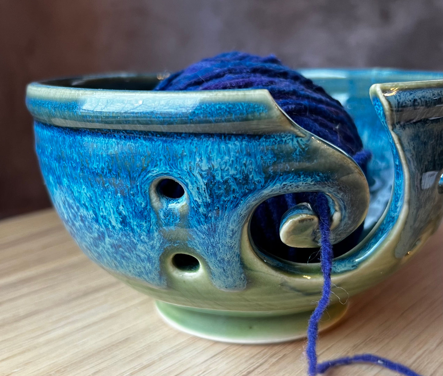 A handmade ceramic yarn bowl with a spiral design, holding a skein of blue yarn, placed on a wooden surface.